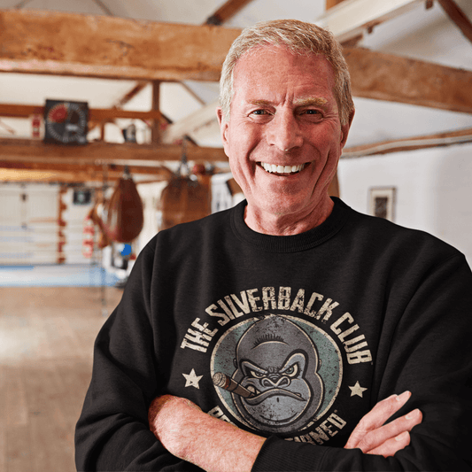 A man wearing a black sweatshirt in a boxing gym with a logo of a silverback gorilla head smoking a cigar and the words The Silverback Club and Respect.Earned surrounding it.