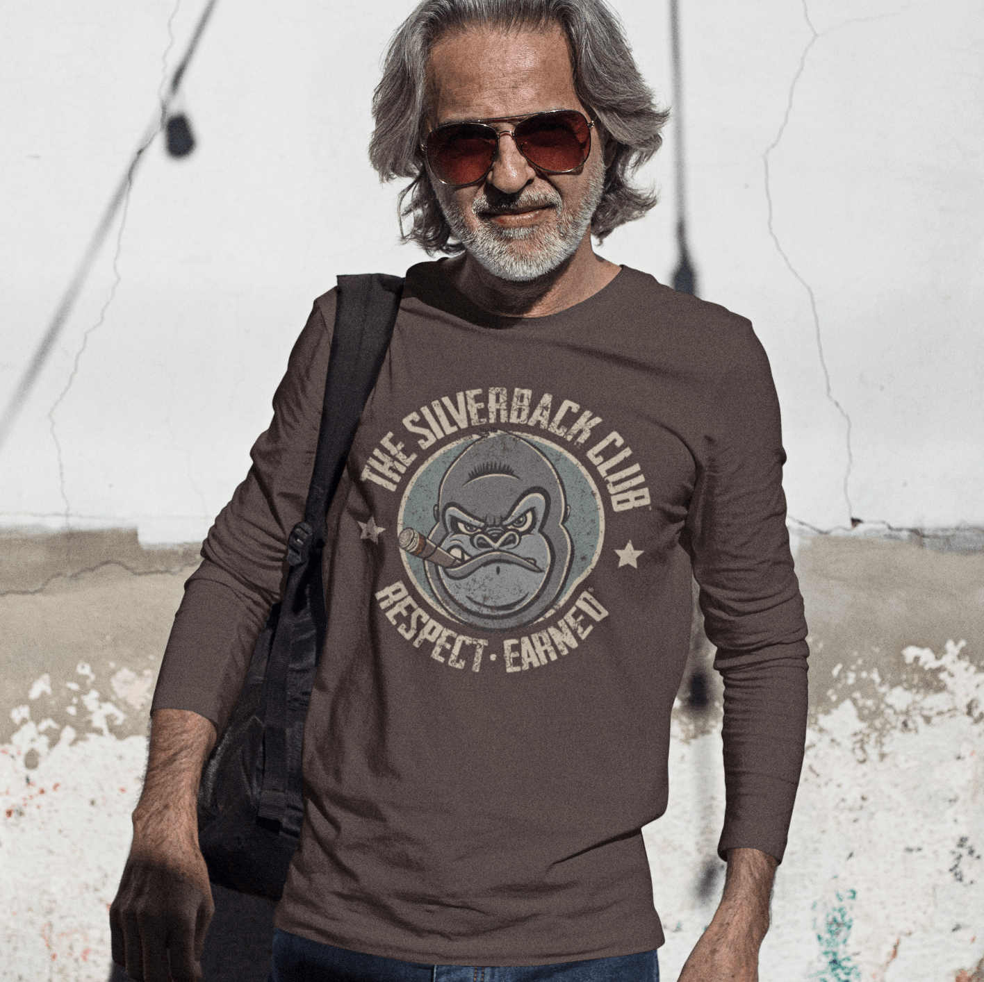 A man with a beard wearing sunglasses and a brown, long-sleeved t-shirt with a logo of a silverback gorilla head smoking a cigar and the words The Silverback Club and Respect.Earned surrounding it.