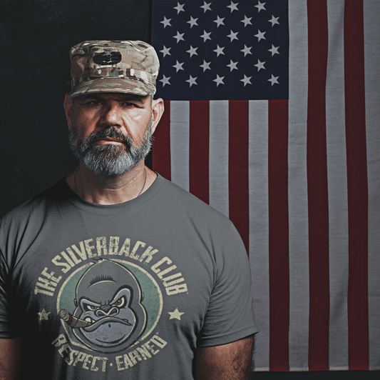 A U.S. military man standing in front of the U.S. flag wearing a grey t-shirt with a logo of a silverback gorilla head smoking a cigar and the words The Silverback Club and Respect.Earned surrounding it.