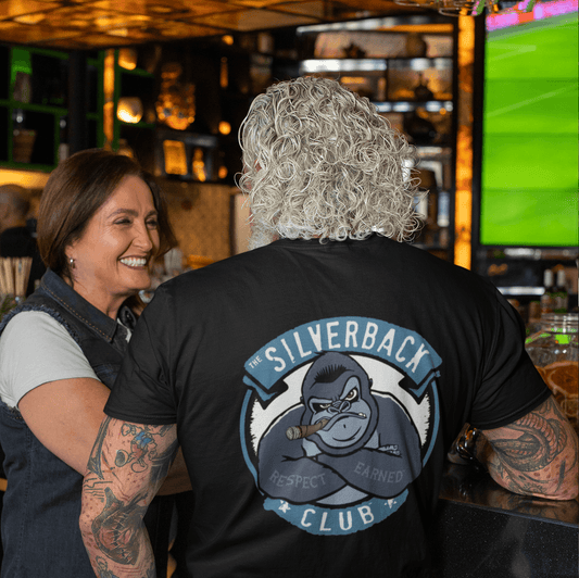 A woman and a man are at a bar and the man has his back to the camera. He is wearing a black t-shirt with a graphic on the back with the words "The Silverback Club" and "Respect" and "Earned" are on a silverback gorillas arms.