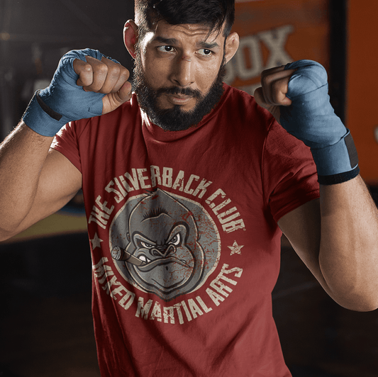 A man wearing a red t-shirt with The Silverback Club logo consisting of a gorilla face smoking a cigar and the words "Mixed Martial Arts" underneath, standing in a fighting pose.