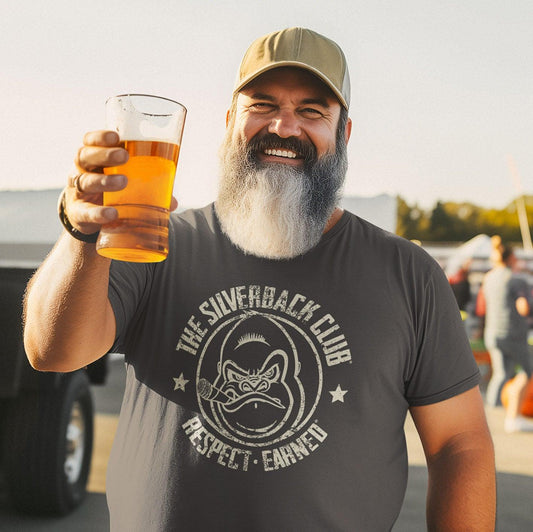 A man wearing a hat, holding a beer is wearing a gray t-shirt with The Silverback Club logo on it of a gorilla smoking a cigar and the words “Respect.Earned” underneath.