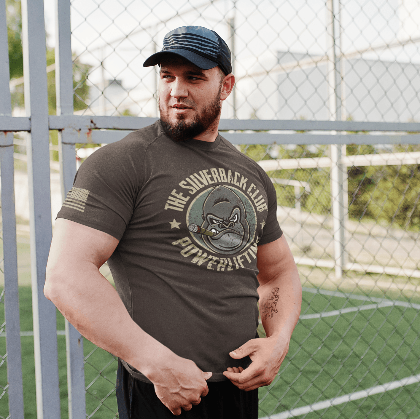 A man wearing a brown t-shirt with The Silverback Club logo consisting of a gorilla face smoking a cigar and the words "Powerlifting" underneath is standing next to a fence.