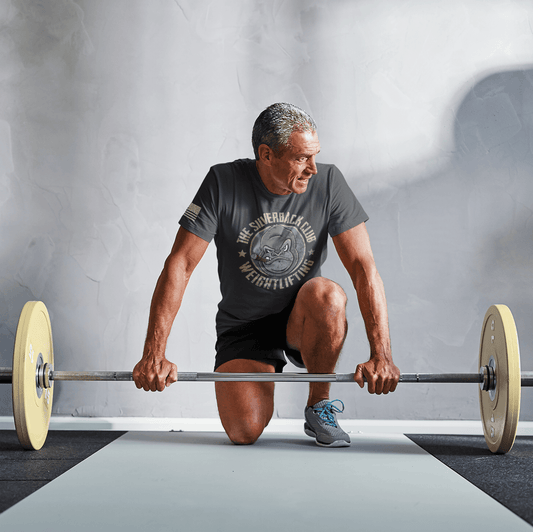 A man wearing a vintage black t-shirt with The Silverback Club logo consisting of a gorilla face smoking a cigar and the words "Weightlifting" underneath is preparing to lift a bar with weights.